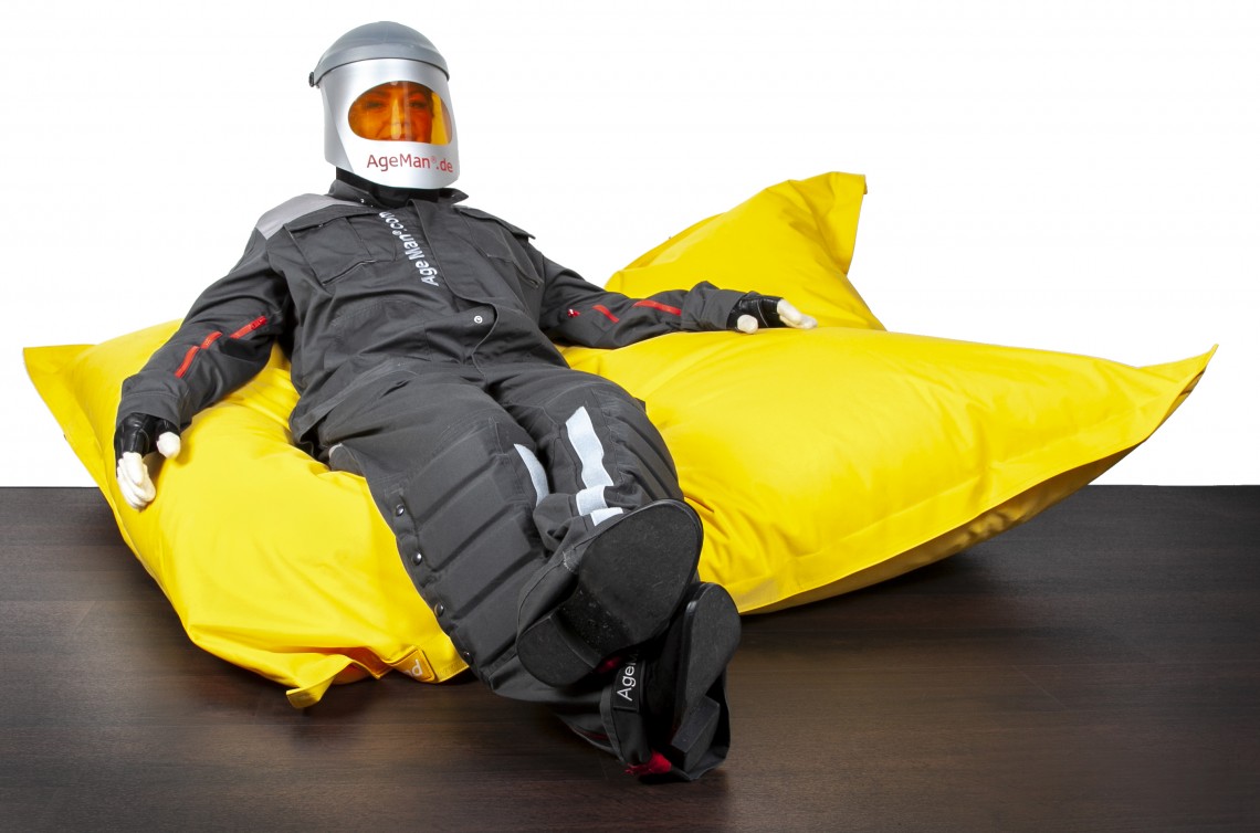 Junge Frau mit angelegtem AgeMan Alterssimulationsanzug liegt in entspannter Position auf einem Fatboy Sitzkissen