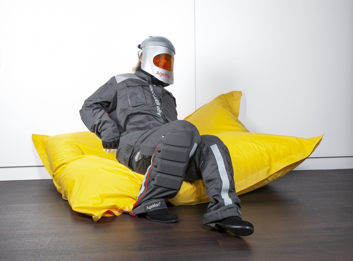 Junge Frau mit angelegtem AgeMan Alterssimulationsanzug beim Aufstehen von einem Fatboy Sitzkissen - Phase 1