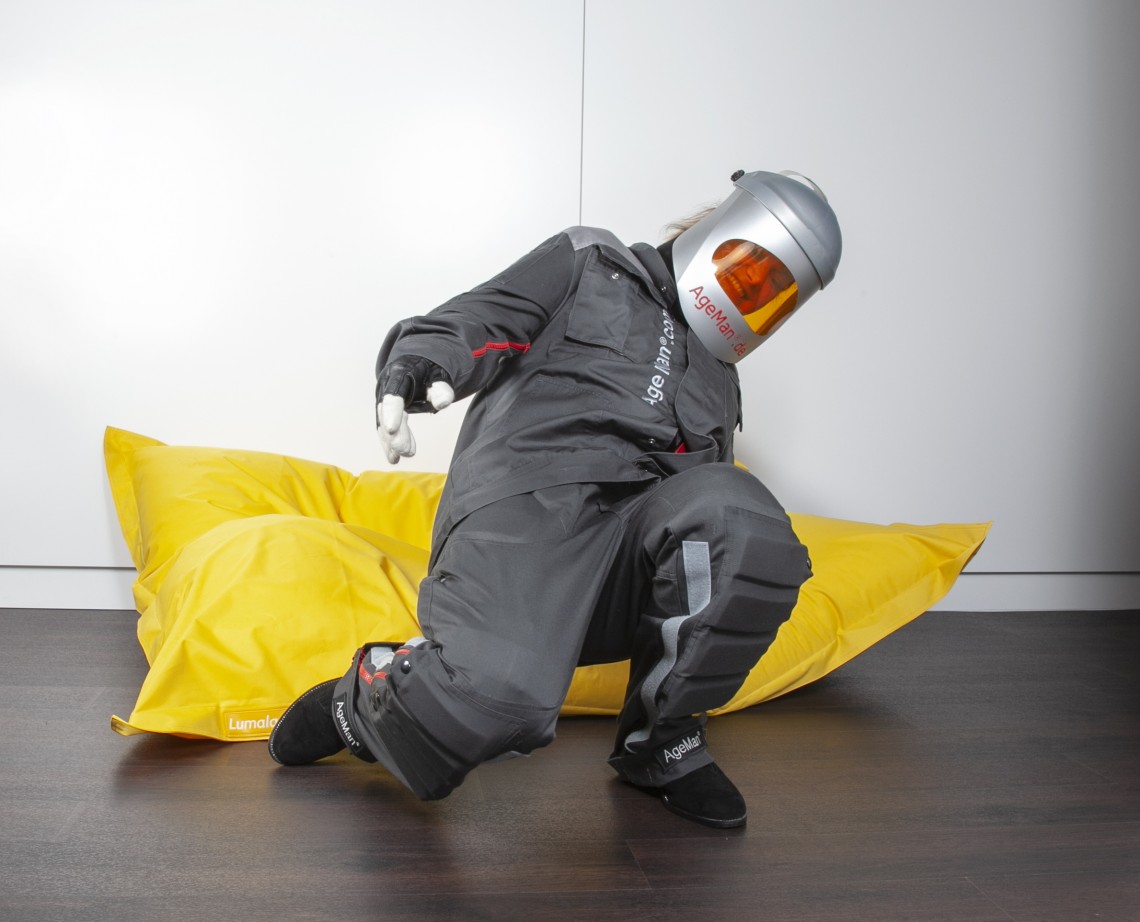 Junge Frau mit angelegtem AgeMan Alterssimulationsanzug beim Aufstehen von einem Fatboy Sitzkissen - Phase 2