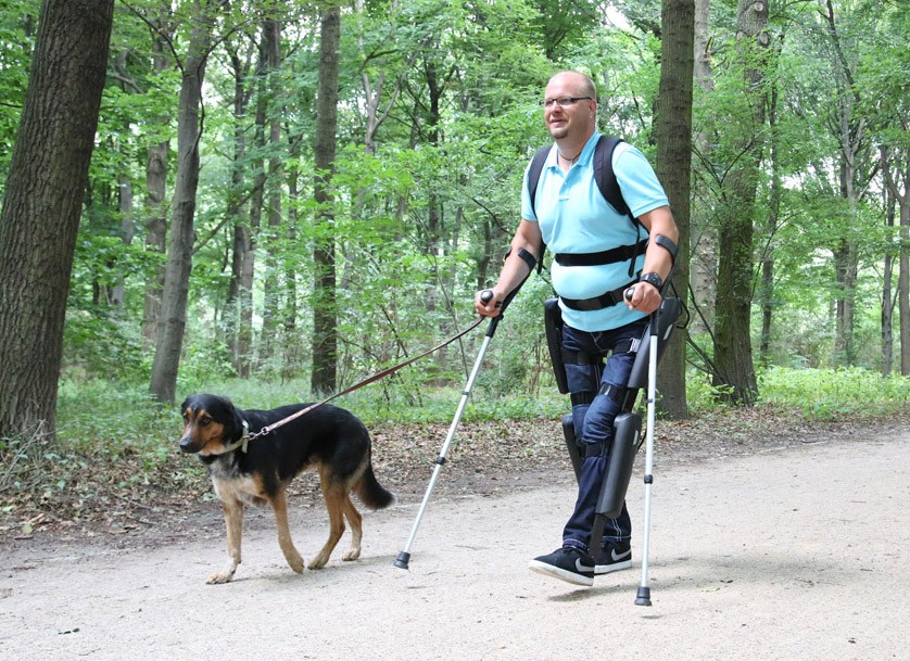 Eine querschnittsgelähmte Person mit angelegtem „ReWalk“ Exoskelett und Krücken auf einem Spaziergang im Wald mit Hund